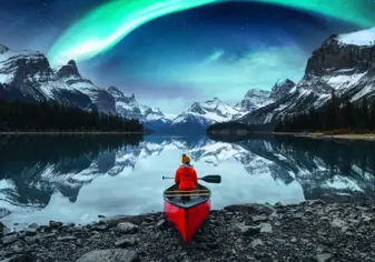 a single person sitting in a canoe at the edge of a lake with mountains in the distance and northern lights in the sky