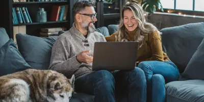 Man and woman sitting on a couch with their dog sipping coffee and looking at tablet