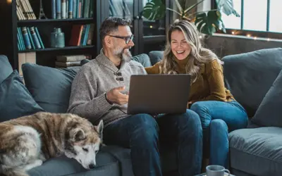 Man and woman sitting on a couch with their dog sipping coffee and looking at tablet